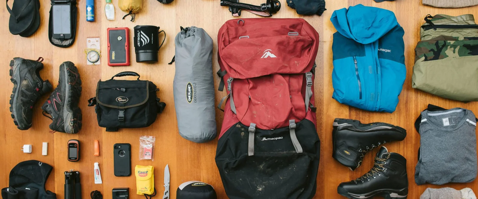 Equipo de trekking en capas para clima frío dispuesto sobre una cama de campamento, incluyendo chaqueta de plumón, forro polar, capa exterior impermeable, guantes y linterna frontal