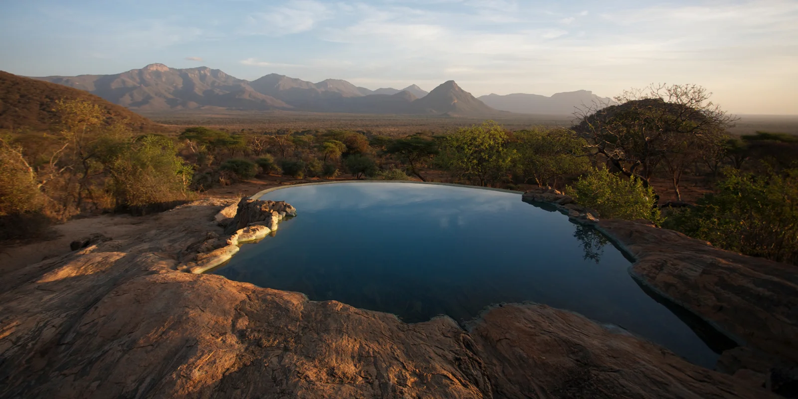 Der Felsbecken-Schwimmbereich von Sarara Camp mit Elefanten, die am angrenzenden Wasserloch trinken